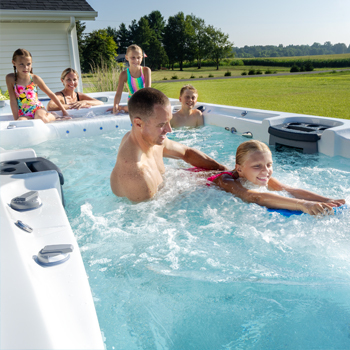 Family enjoying time together in a swim spa and a man helping his daughter on a kickboard.